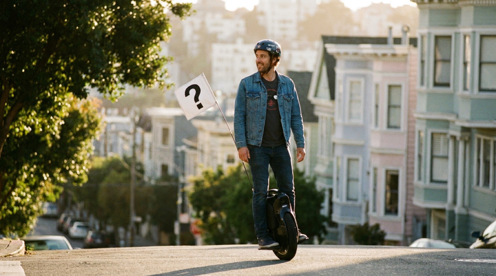 Rider on a black electric unicycle descending a San Francisco street at golden hour, a blank white flag on a thin whip rising from the back of the wheel, cable car and Victorian houses behind